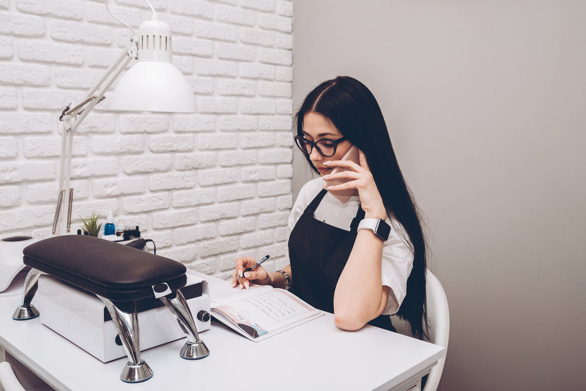 Nail technician wearing glasses and an apron, talking on the phone while checking her appointment book at a salon desk.